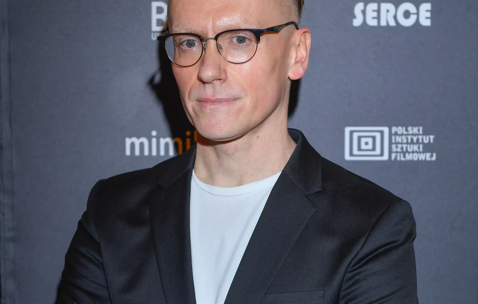 Mężczyzna w okularach, ubrany w czarną marynarkę i białą koszulkę, stoi na tle ścianki z logo Polskiego Instytutu Sztuki Filmowej.