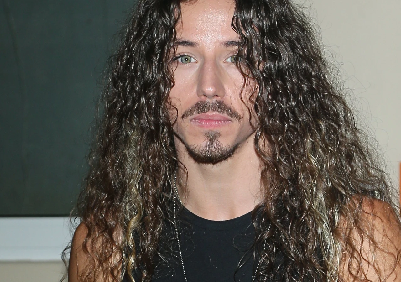 Michał Szpak Michał Szpak z długimi, kręconymi włosami i zarostem, ubrany w czarną koszulkę, patrzy w obiektyw aparatu.