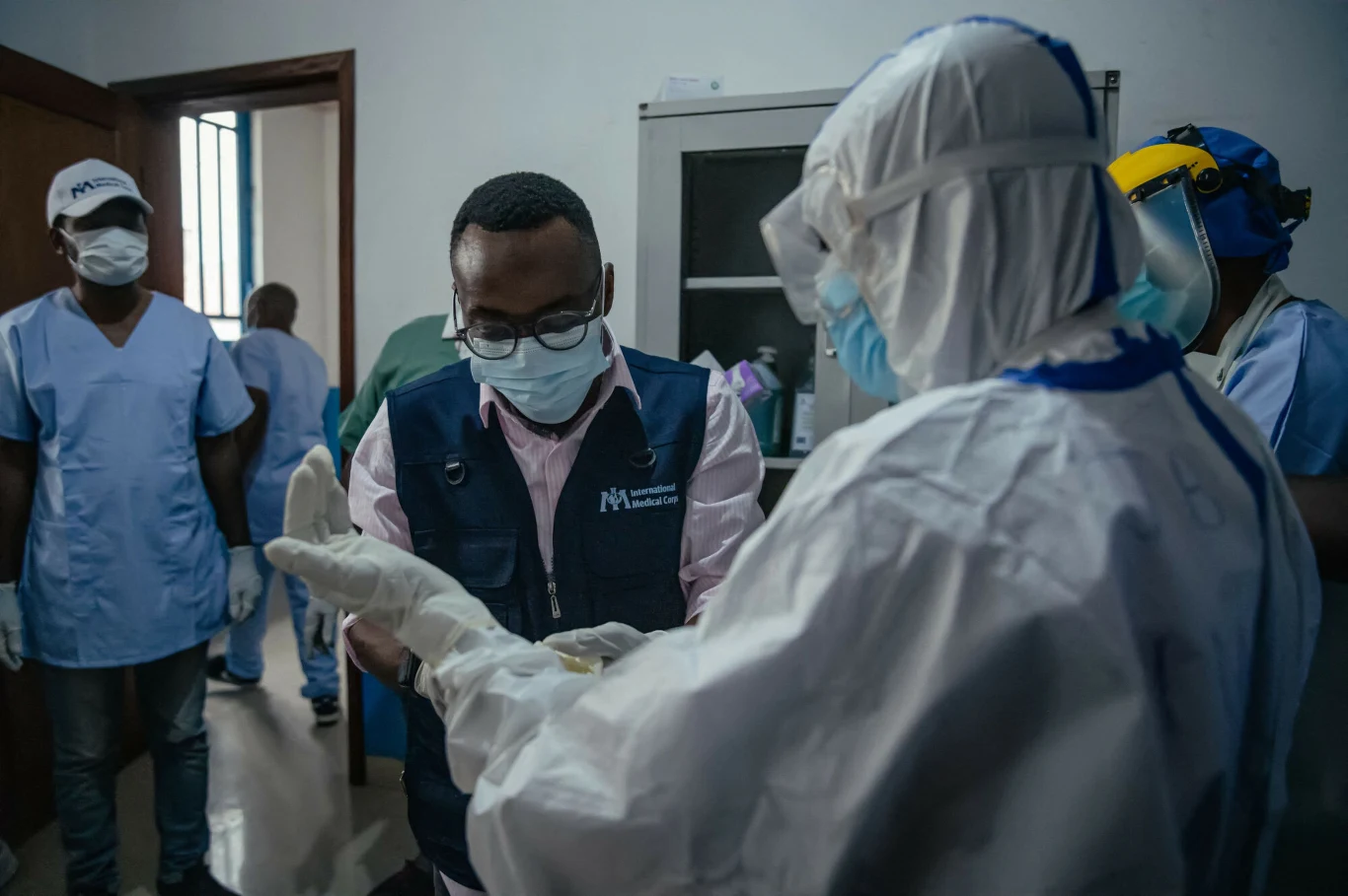 Ebolę da się kontrolować - mówią eksperci Lekarz w okularach i maseczce rozmawia z osobą w pełnym stroju ochronnym, w tle kilku pracowników medycznych w ubraniach roboczych.