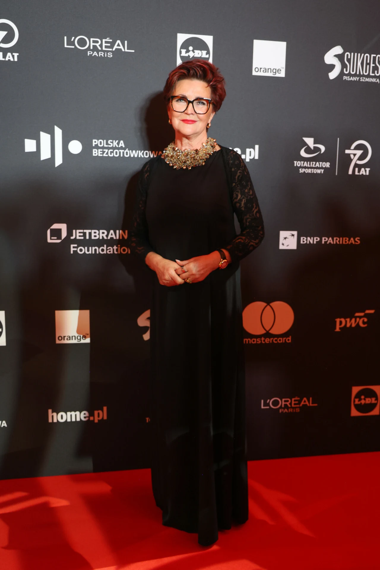 Elegancka kobieta w czarnej długiej sukni z koronkowymi rękawami i efektowną ozdobną kolią, stojąca na czerwonym dywanie na tle ściany z logo sponsorów.