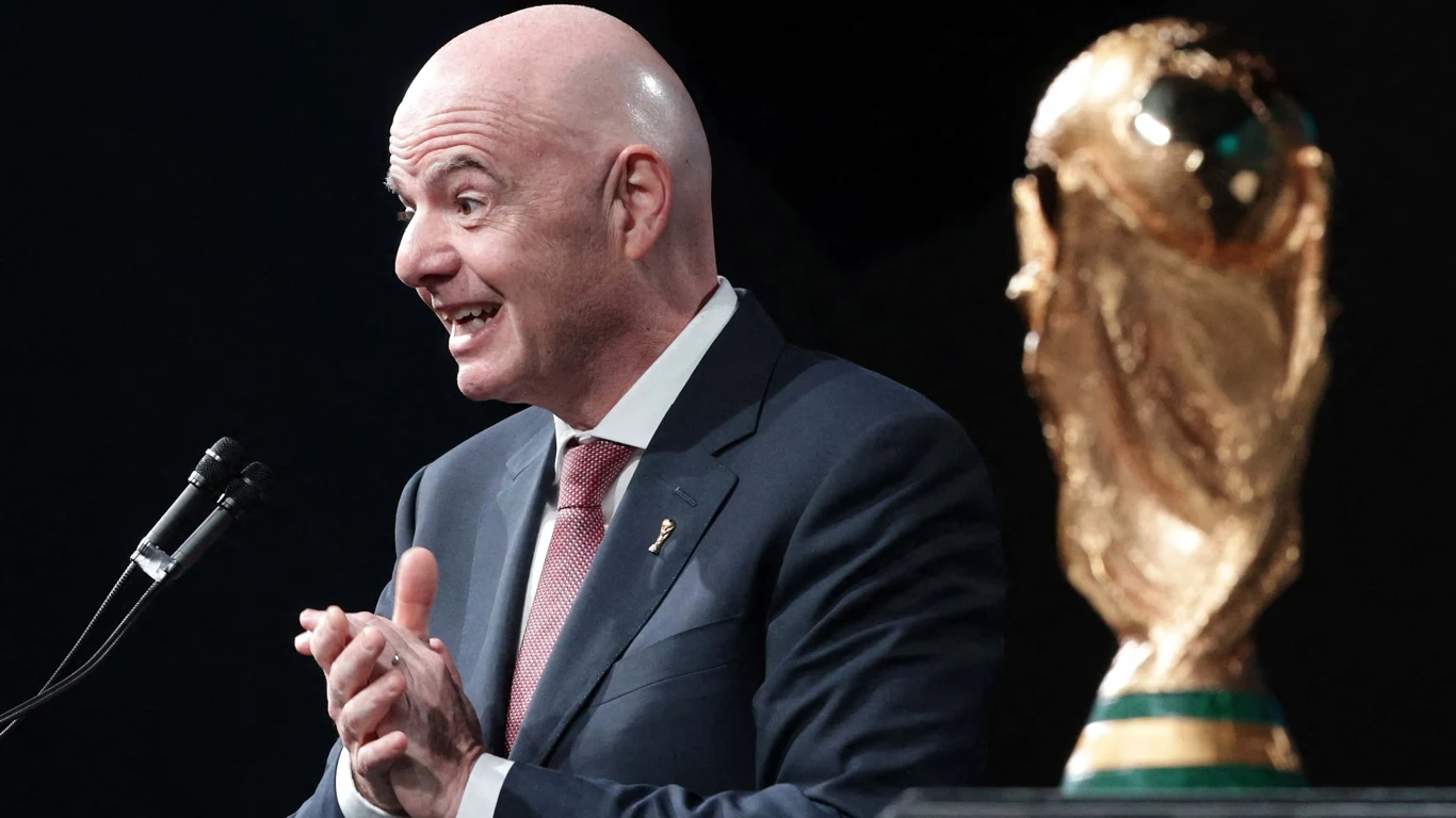 Łysy mężczyzna w garniturze przemawia do mikrofonu, obok złoty puchar Mistrzostw Świata FIFA.