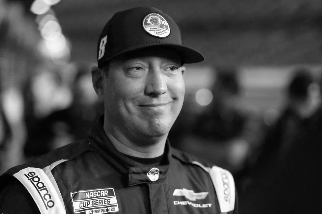 Uśmiechnięty kierowca wyścigowy w czapce z daszkiem i kombinezonie z naszywkami sponsorów, w tym logo NASCAR Cup Series i Chevrolet.