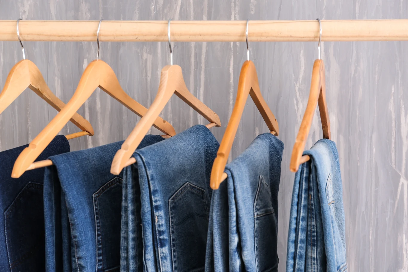 Modny i uniwersalny fason jeansów. Złoty środek pomiędzy wąskimi rurkami, a szerokimi spodniami oversize. Kilka par jeansów stovepipe w różnych odcieniach niebieskiego na drewnianych wieszakach na metalowym drążku.