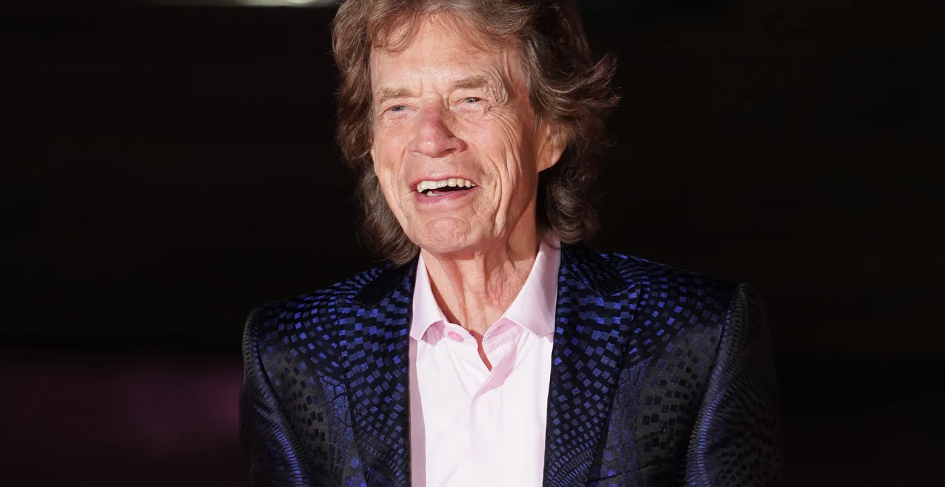 Mick Jagger Mick Jagger w eleganckiej marynarce we wzory i jasnoróżowej koszuli na ciemnym tle.