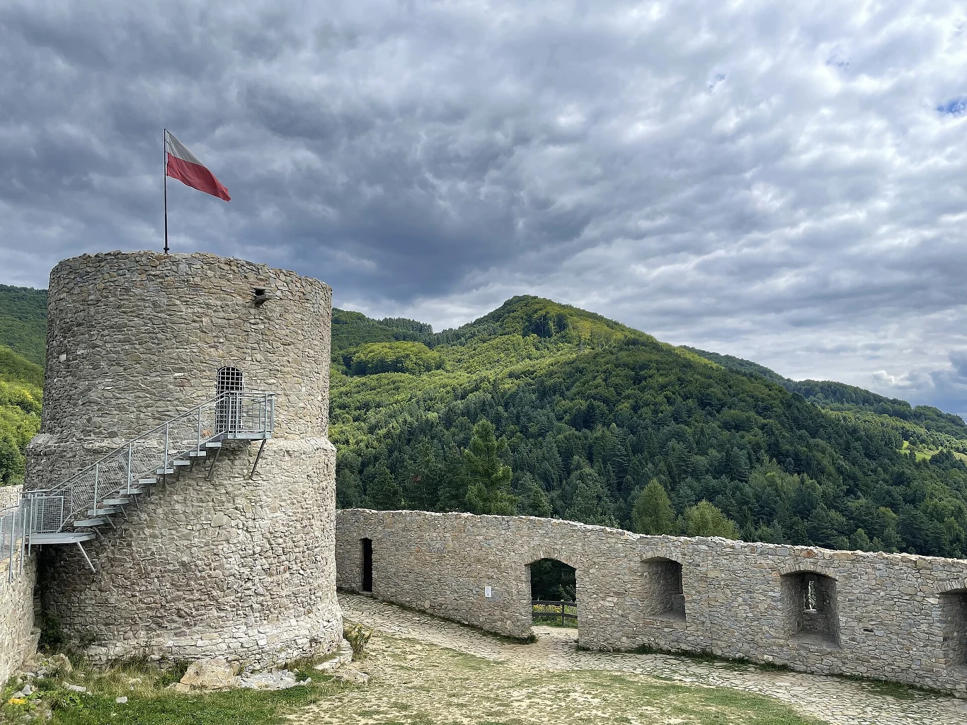 Kamienna wieża z polską flagą na szczycie, fragment murów obronnych oraz rozległe, zielone wzgórza w tle.