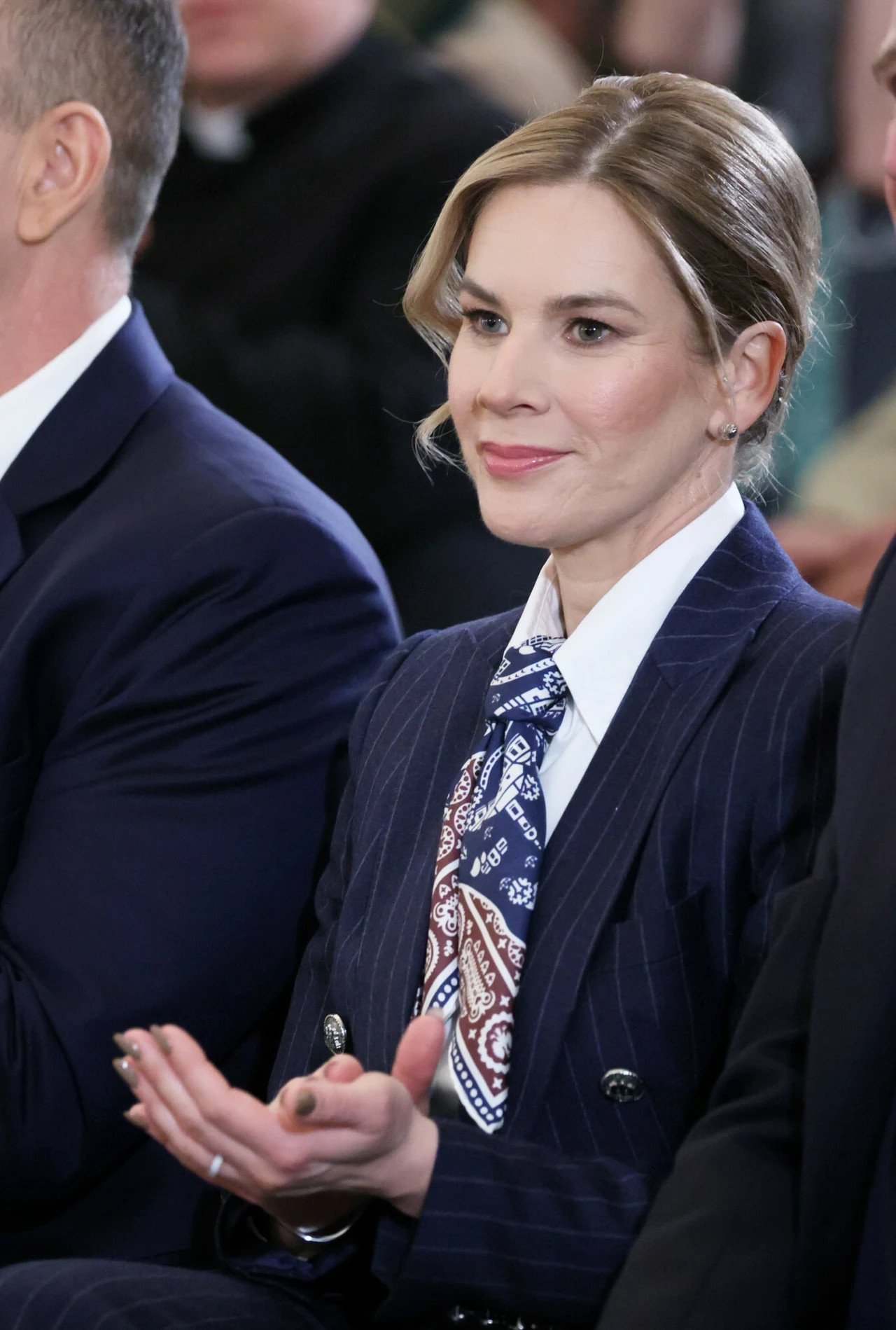 Apaszka sprawdzi się też przy formalnych i bardzo eleganckich outfitach Kobieta w eleganckim garniturze w prążki i wzorzystej apaszce siedzi wśród innych osób, z delikatnym uśmiechem i dłońmi położonymi na kolanach.