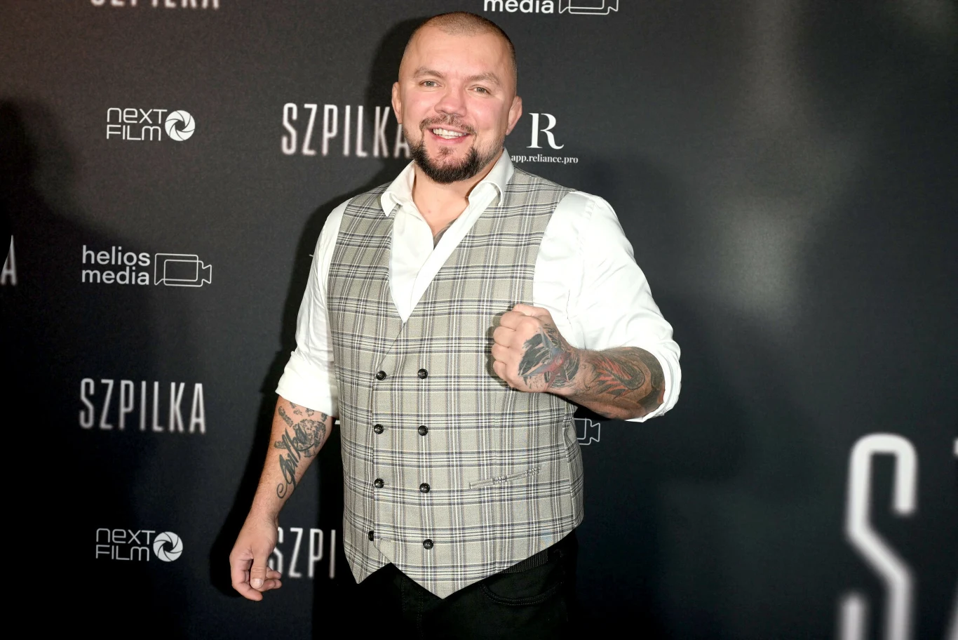 Łukasz "Juras" Jurkowski Uśmiechnięty mężczyzna w jasnej koszuli i szarej kamizelce w kratę z tatuażami na rękach pozuje do zdjęcia na tle eventowej ścianki z czarnym tłem i białymi napisami.