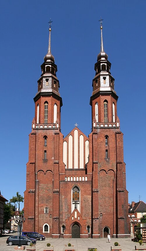 Ceglany kościół z dwoma wysokimi wieżami i strzelistymi hełmami, położony przy brukowanym placu z zaparkowanymi samochodami.