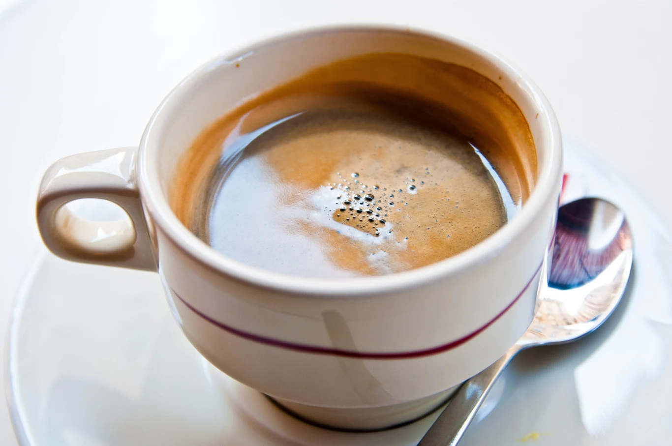 Ta przyprawa dodawana codziennie do kawy pomoże ci uregulować poziom cukru we krwi. Biała filiżanka espresso na spodku, obok leży łyżeczka; ilustracja kawy z kardamonem wspierającej poziom cukru.