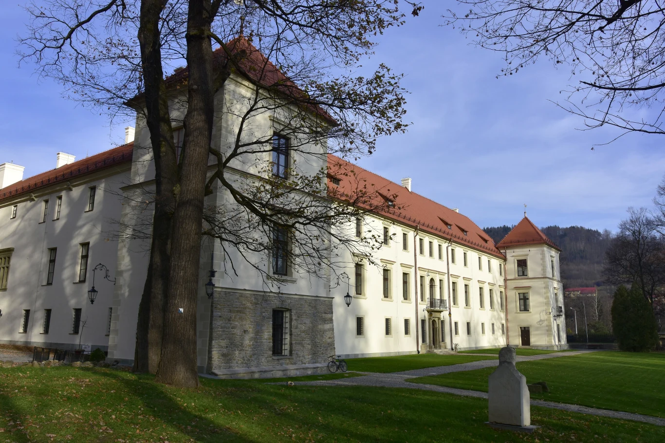 Renesansowy pałac z wysokimi wieżami, jasną elewacją i czerwonym dachem otoczony starannie przystrzyżonym trawnikiem oraz pojedynczymi drzewami w bliskim sąsiedztwie.