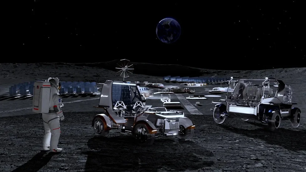 Astronauta w skafandrze kosmicznym spogląda na dwa zaawansowane łaziki stojące na powierzchni Księżyca, w tle widoczne zabudowania bazy księżycowej oraz Ziemia na czarnym niebie.