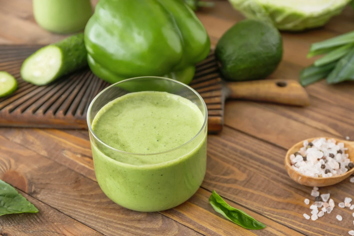 Szklanka kremowego zielonego smoothie na drewnianym stole, otoczona awokado, papryką i ogórkiem, z dodatkiem gruboziarnistej soli morskiej w miseczce.