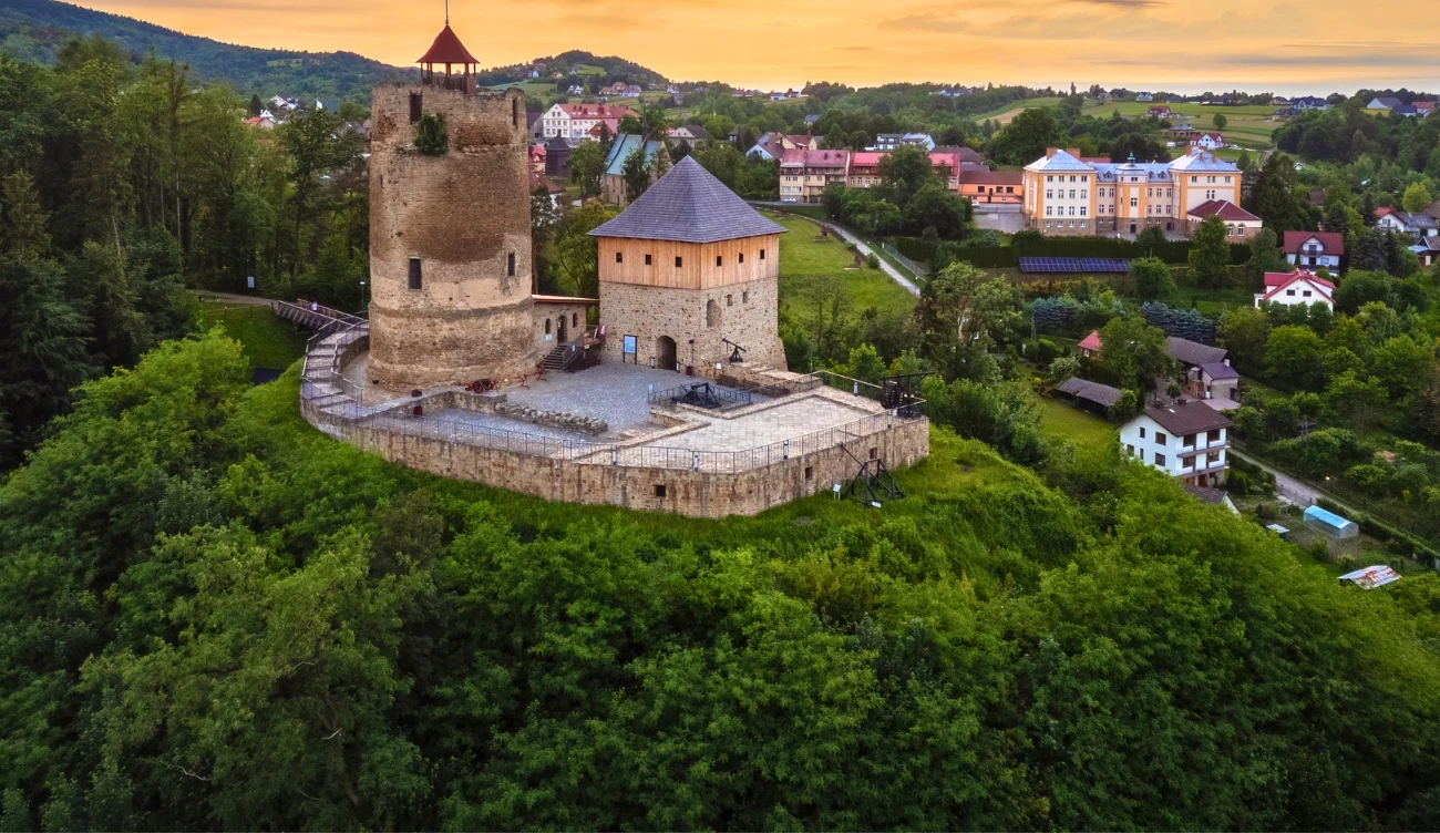 Cylindryczna wieża w Czchowie Kamienna wieża zamku z czterospadowym dachem otoczona murami obronnymi, położona na wzgórzu porośniętym gęstą zielenią, z widoczną w tle zabudową miasteczka.