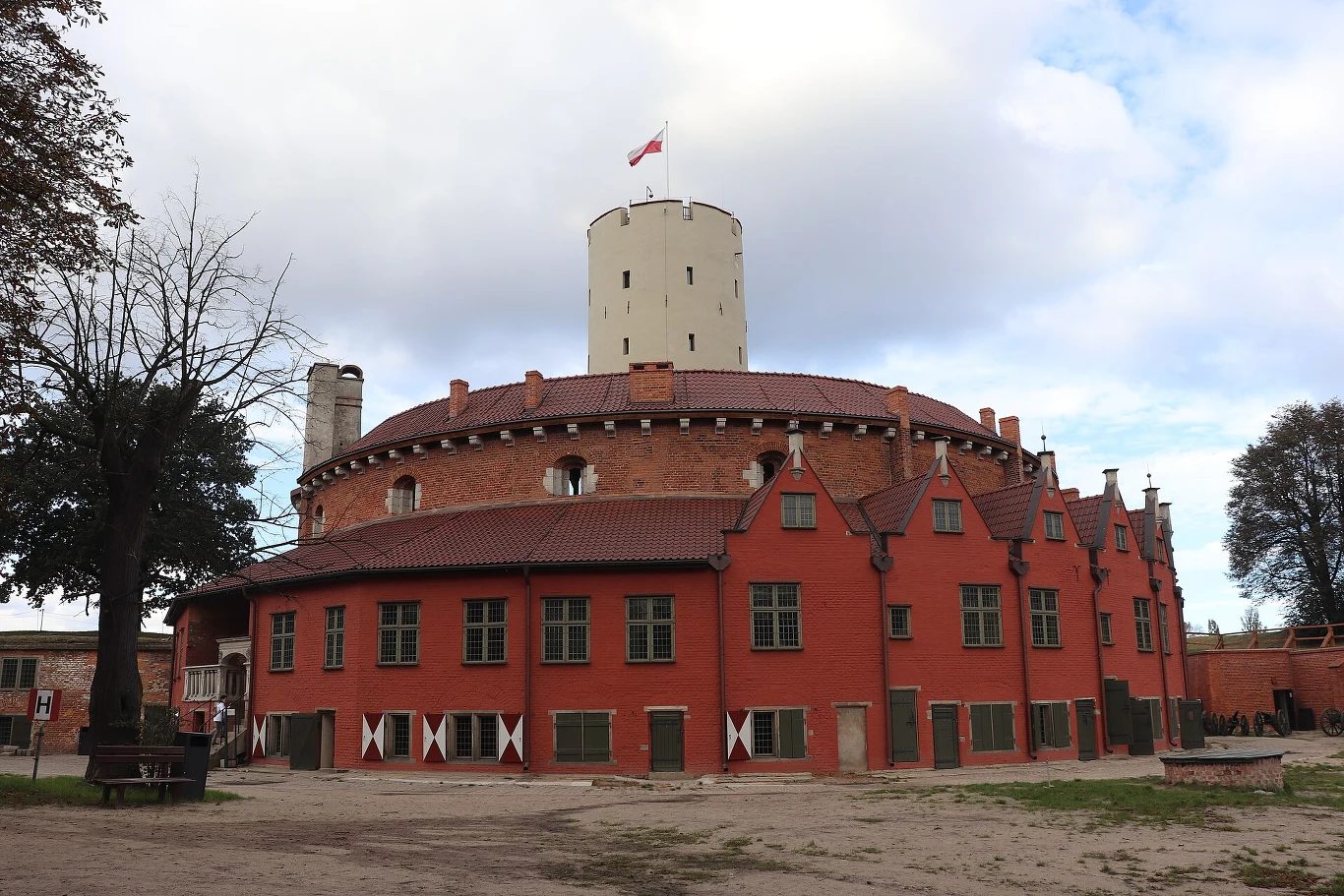 Ceglana Twierdza Wisłoujście, okrągła wieża z polską flagą na szczycie, otoczona drzewami i dziedzińcem