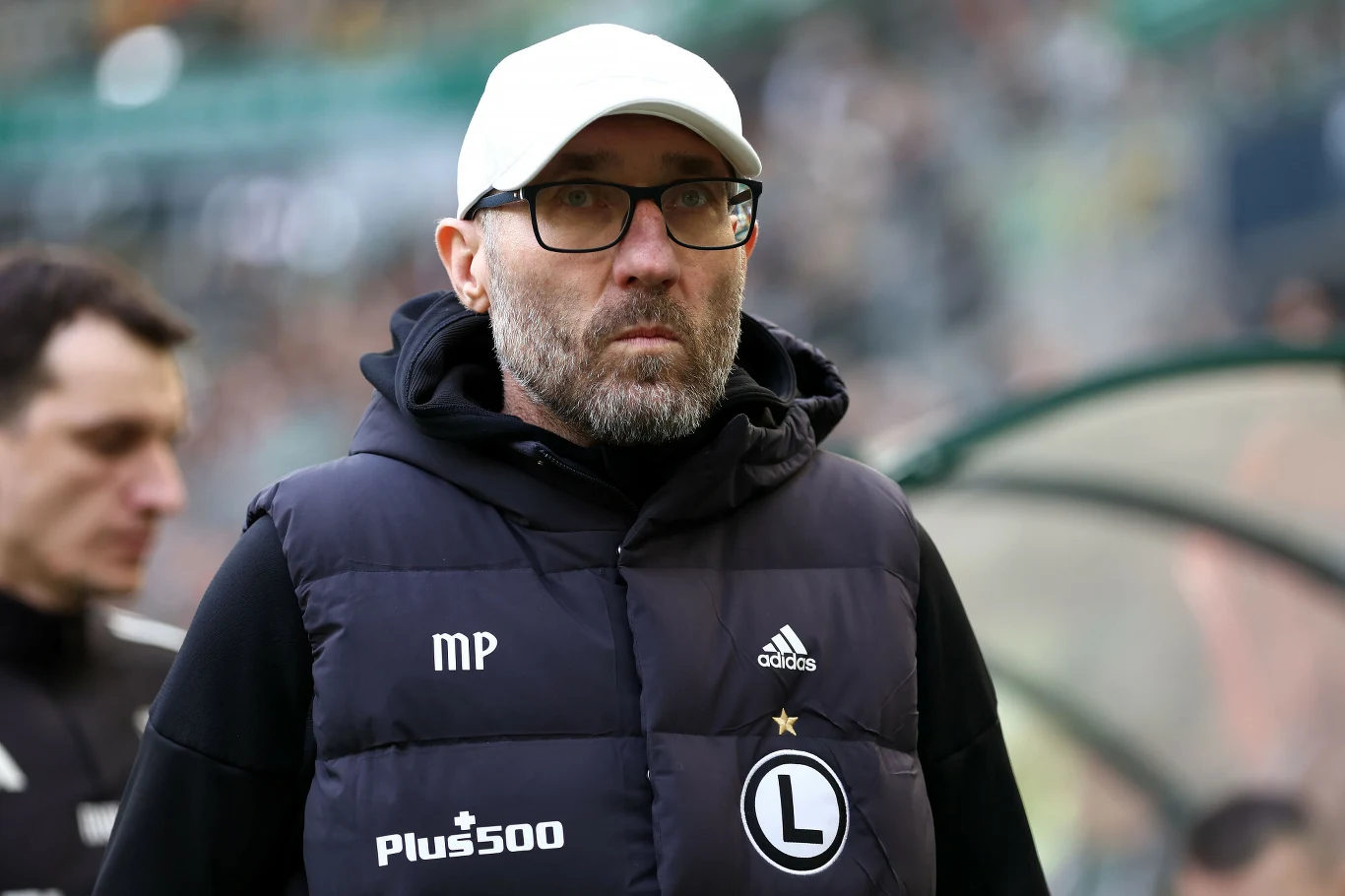 Mężczyzna w okularach i białej czapce z daszkiem ubrany w ciemną kamizelkę sportową z logo Legii Warszawa oraz naszywkami Adidas i Plus500, stojący na stadionie piłkarskim.
