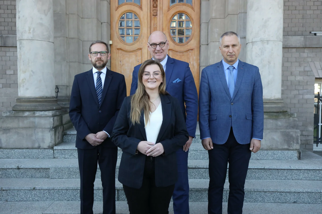 Cztery osoby w eleganckich garniturach stojące na schodach przed wejściem do dużego budynku z drewnianymi drzwiami i masywnymi kolumnami.