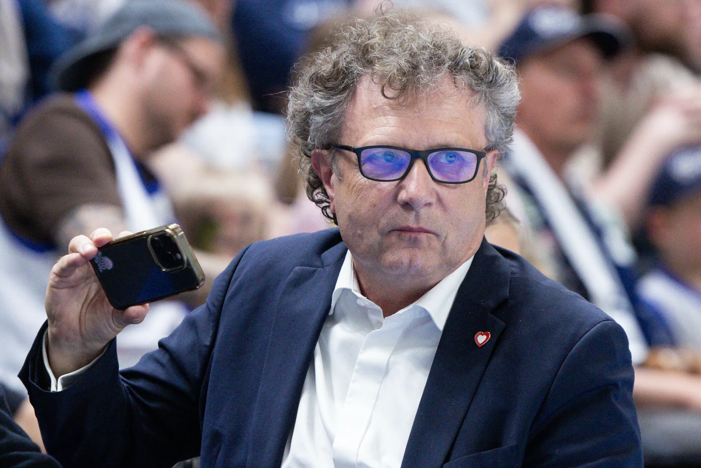 Mężczyzna w okularach i granatowej marynarce trzyma telefon z uniesioną ręką, w tle rozmyta publiczność.