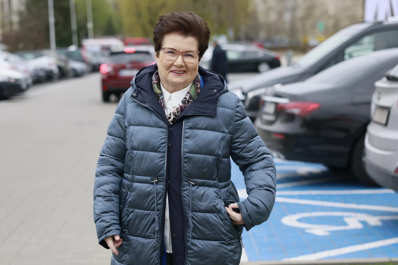 Uśmiechnięta kobieta w okularach i pikowanej kurtce spaceruje po parkingu, stojąc na miejscu dla osób z niepełnosprawnościami.