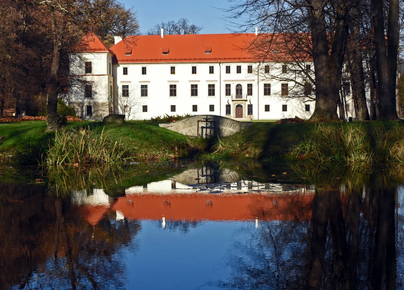 Duży biały pałac z czerwonym dachem, stojący nad stawem, w którym odbija się jego fasada, otoczony drzewami w jesiennych barwach.