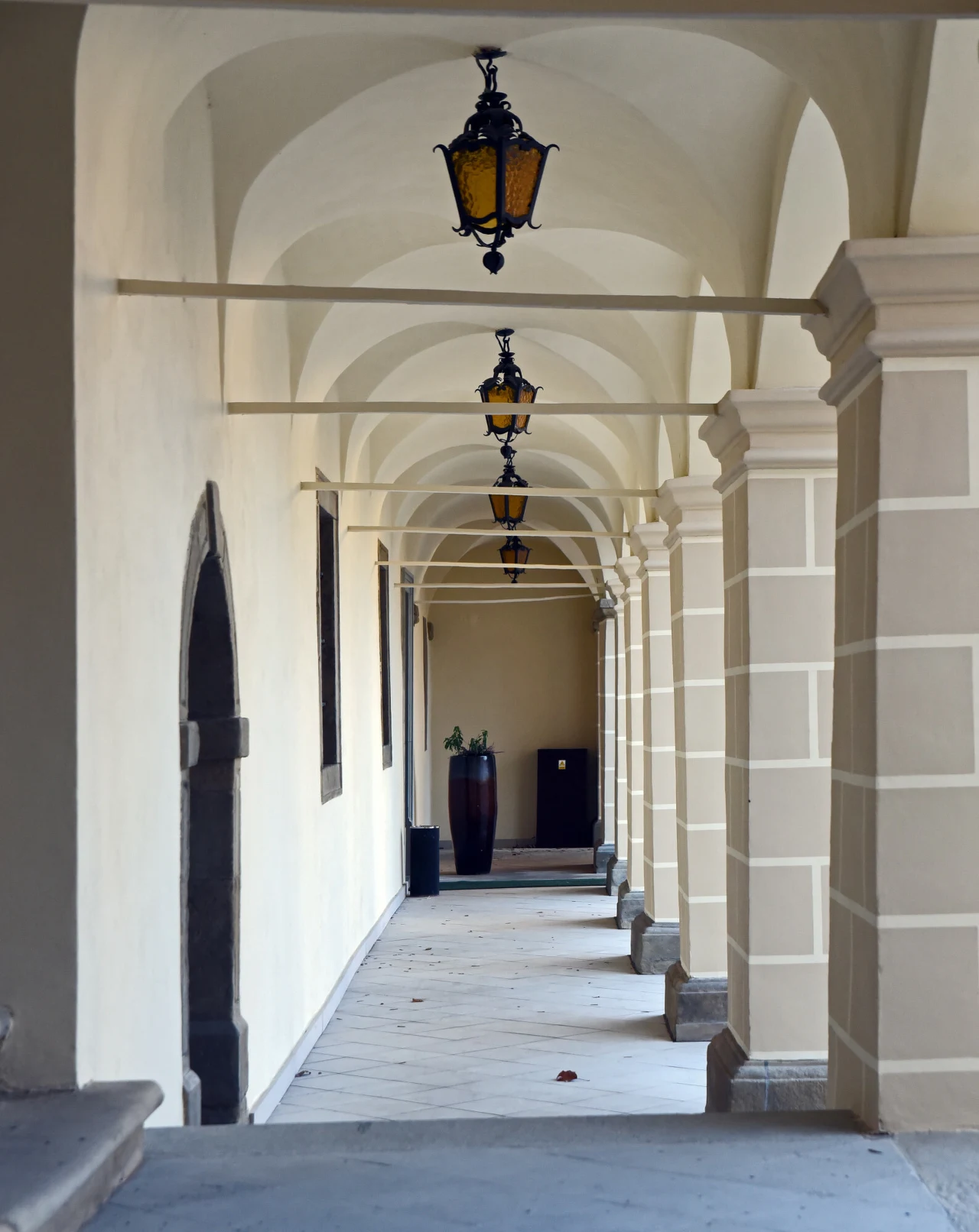 Zabytkowy korytarz z rzędem łukowych sklepień i ozdobnych lamp wiszących, prowadzący w głąb budynku, wykończony eleganckimi kolumnami oraz wazonem z zieloną rośliną na końcu przejścia.