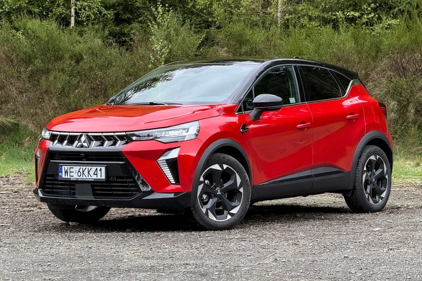 Czerwony samochód typu SUV z czarnym dachem, charakterystycznym przodem z dużym grillem oraz nowoczesnym wzorem reflektorów, stojący na asfaltowej nawierzchni na tle zielonych krzewów.