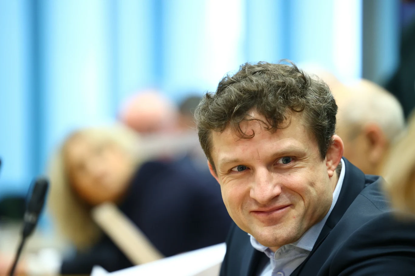 Mężczyzna w garniturze z uśmiechem na twarzy siedzi przy stole, otoczony innymi ludźmi podczas spotkania.
