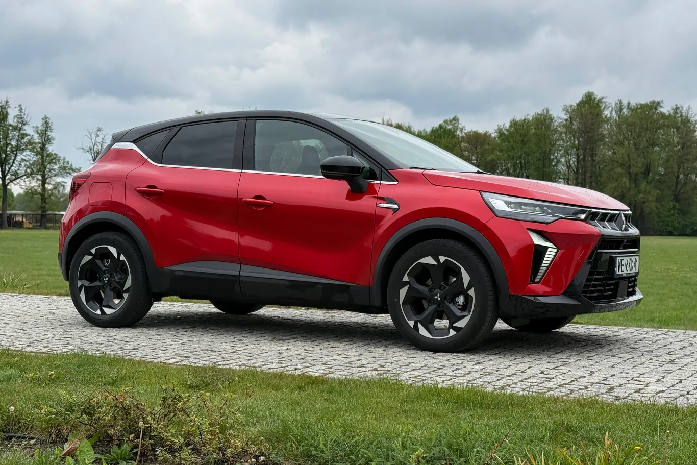 Czerwony, nowoczesny SUV z czarnymi detalami i dużymi felgami stoi na ścieżce z kostki brukowej pośród zielonej trawy; w tle drzewa i pochmurne niebo.