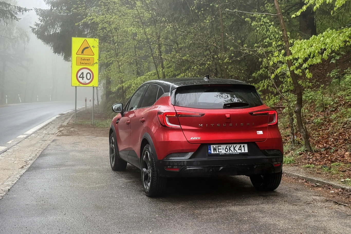 Czerwony samochód osobowy marki Mitsubishi zaparkowany na poboczu leśnej drogi przy znaku ostrzegającym o stromym zjeździe i ograniczeniu prędkości do 40 km/h.