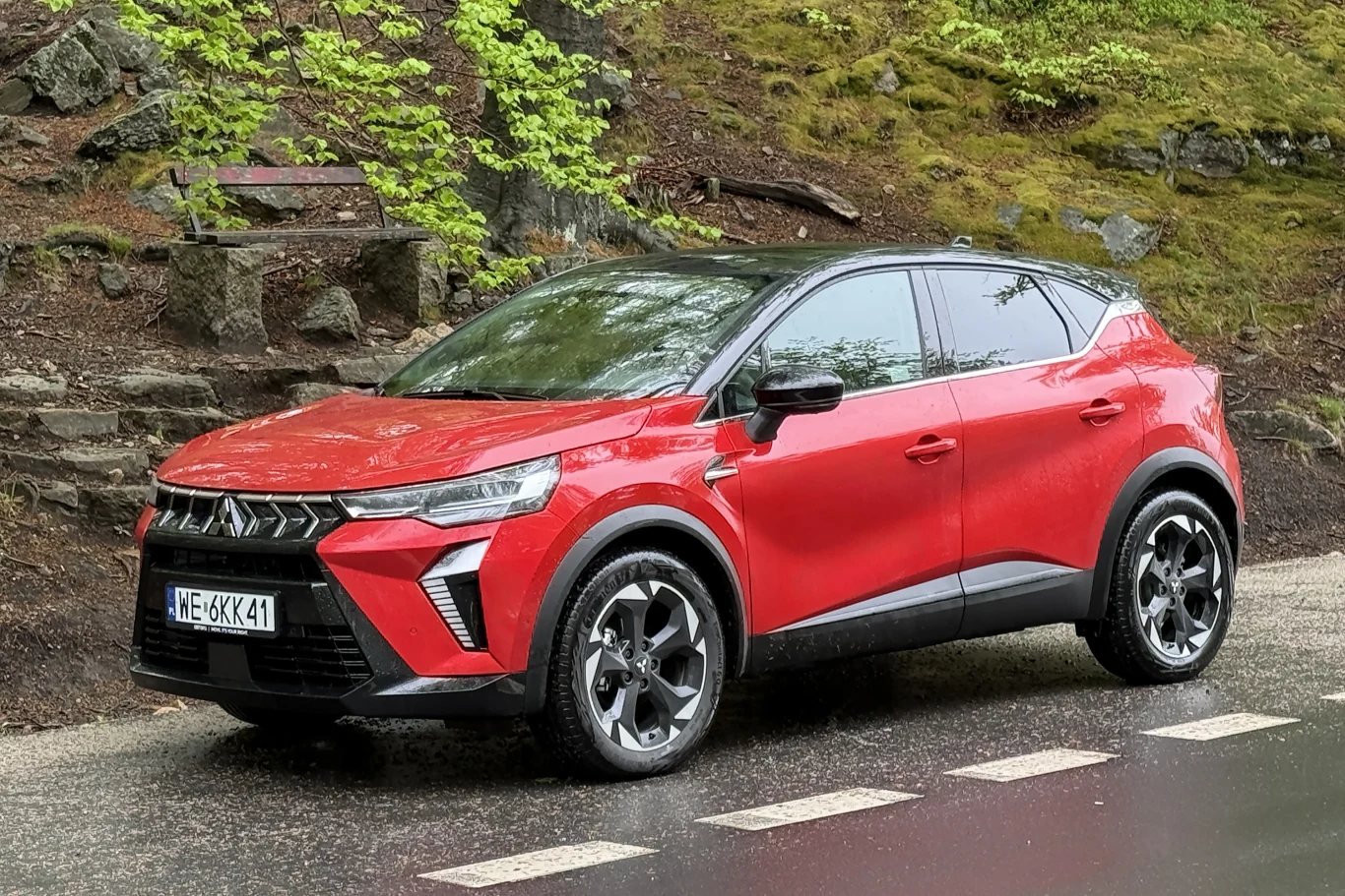 Czerwony SUV Mitsubishi z czarnym dachem stoi zaparkowany przy mokrej ulicy, otoczony zielenią i kamiennym murem.