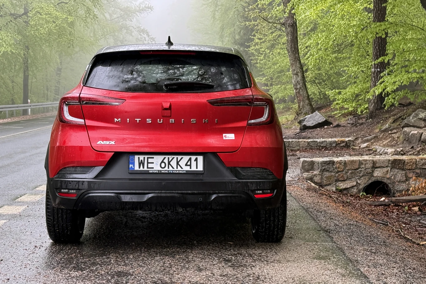 Czerwony samochód Mitsubishi ASX zaparkowany tyłem przy drodze otoczonej zielonymi drzewami, obok kamiennego murka.