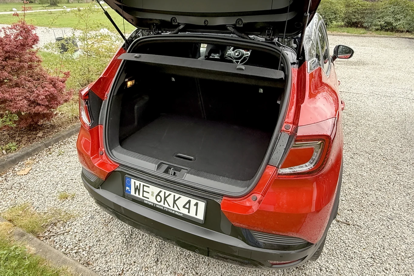 Otwarty bagażnik czerwonego samochodu osobowego, tablica rejestracyjna WE 6KK41, auto stoi na żwirowym placu obok roślinności.