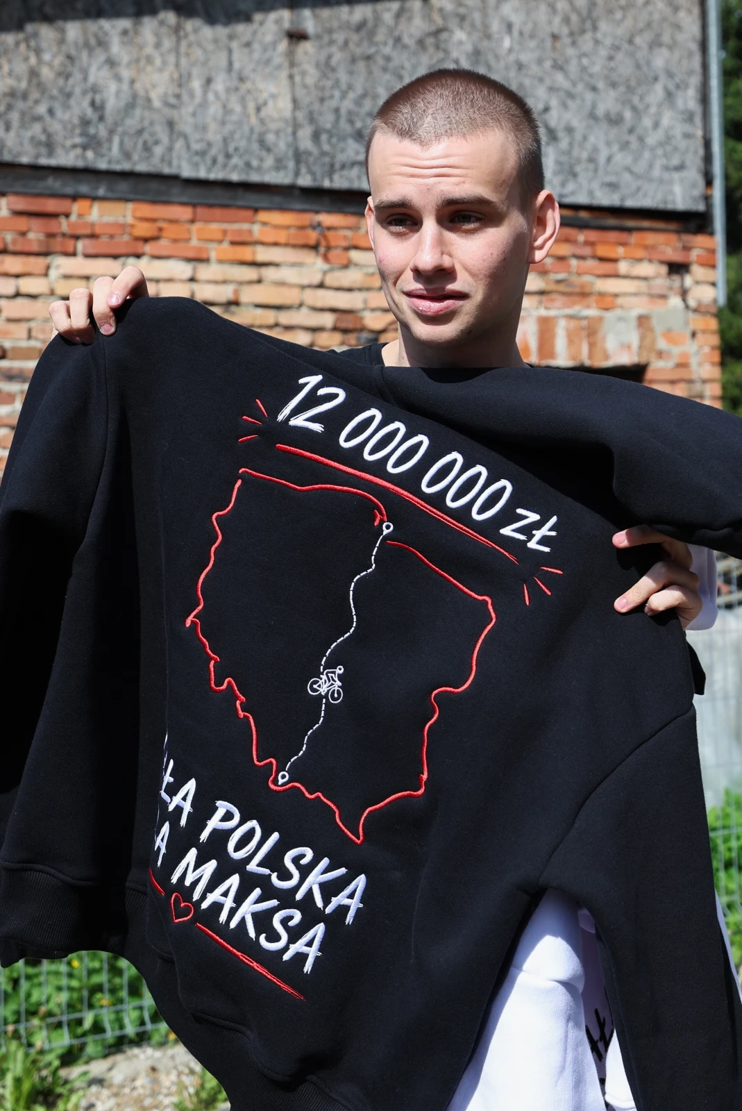 Młody mężczyzna z krótkimi włosami trzyma dużą czarną bluzę z nadrukowaną mapą Polski, napisem „12 000 000 zł” oraz napisem „Dla Polska na maksa”, na tle ceglanej ściany.