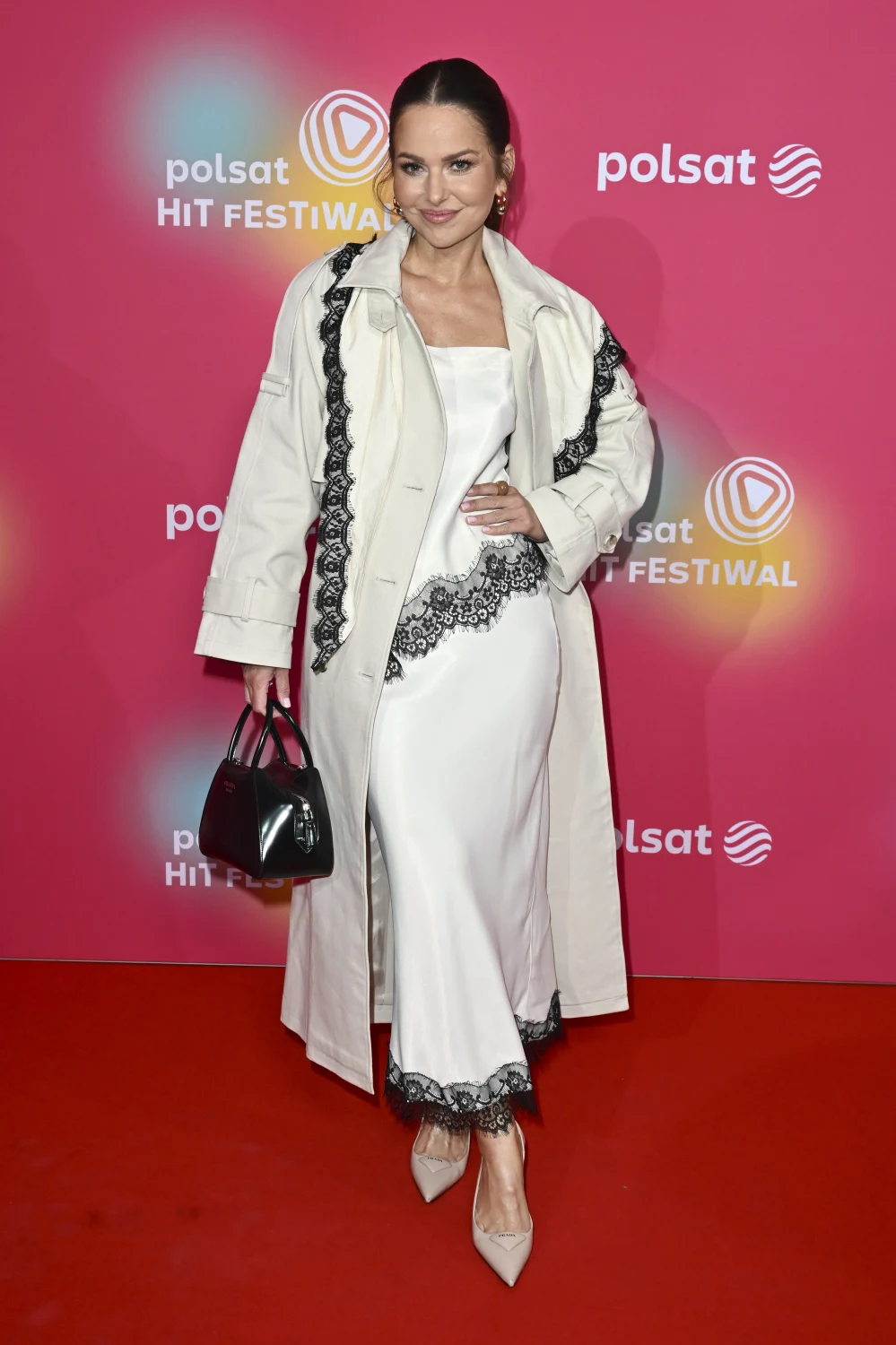 Paulina Sykut-Jeżyna na Polsat Hit Festiwal 2026 Kobieta w długim białym płaszczu z czarnymi zdobieniami i białej sukience z koronką, trzymająca czarną torebkę i pozująca na czerwonym dywanie, w tle ścianka z logo Polsat Hit Festiwal.