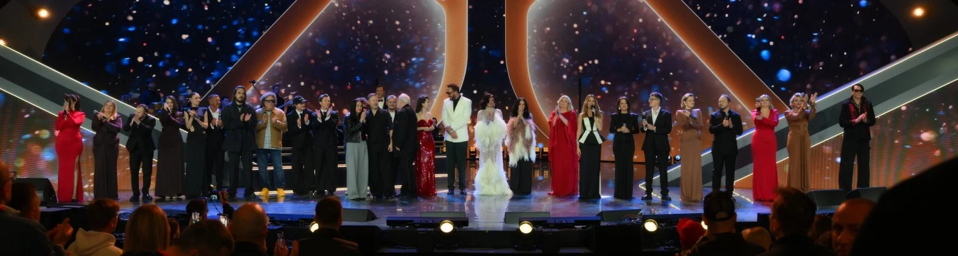 Artyści na koncercie dla Krzysztofa Krawczyka na Polsat Hit Festiwal 2026 Grupa elegancko ubranych osób stojących na scenie udekorowanej efektownym oświetleniem i kolorowymi światłami, wszyscy zwróceni w stronę publiczności i klaszczących widzów.