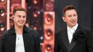 Dwóch mężczyzn ubranych elegancko stoi obok siebie na tle rozświetlonej sceny z dużymi głośnikami, obaj mają na uszach słuchawki bezprzewodowe i wyrażają pozytywne emocje.
