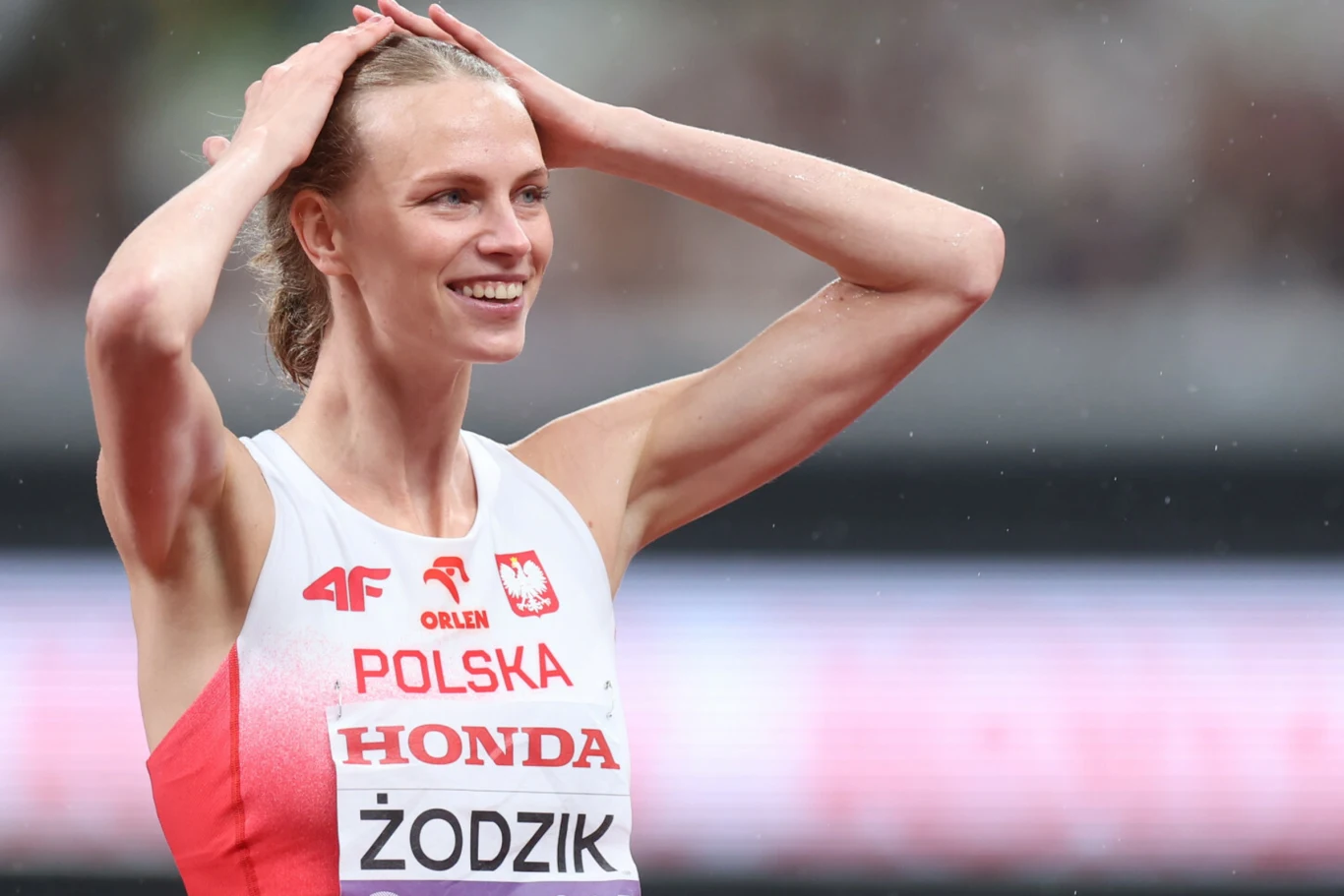Maria Żodzik Zawodniczka w stroju reprezentacji Polski z uśmiechem i rękami na głowie podczas zawodów lekkoatletycznych.