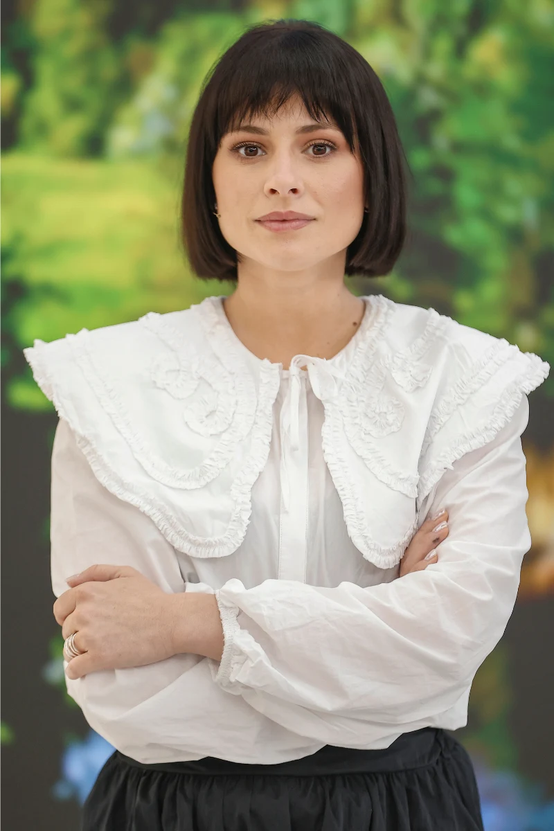 Kobieta z ciemnymi włosami do ramion w białej bluzce z szerokim kołnierzem, stoi z założonymi rękami na tle rozmytego zielonego krajobrazu.