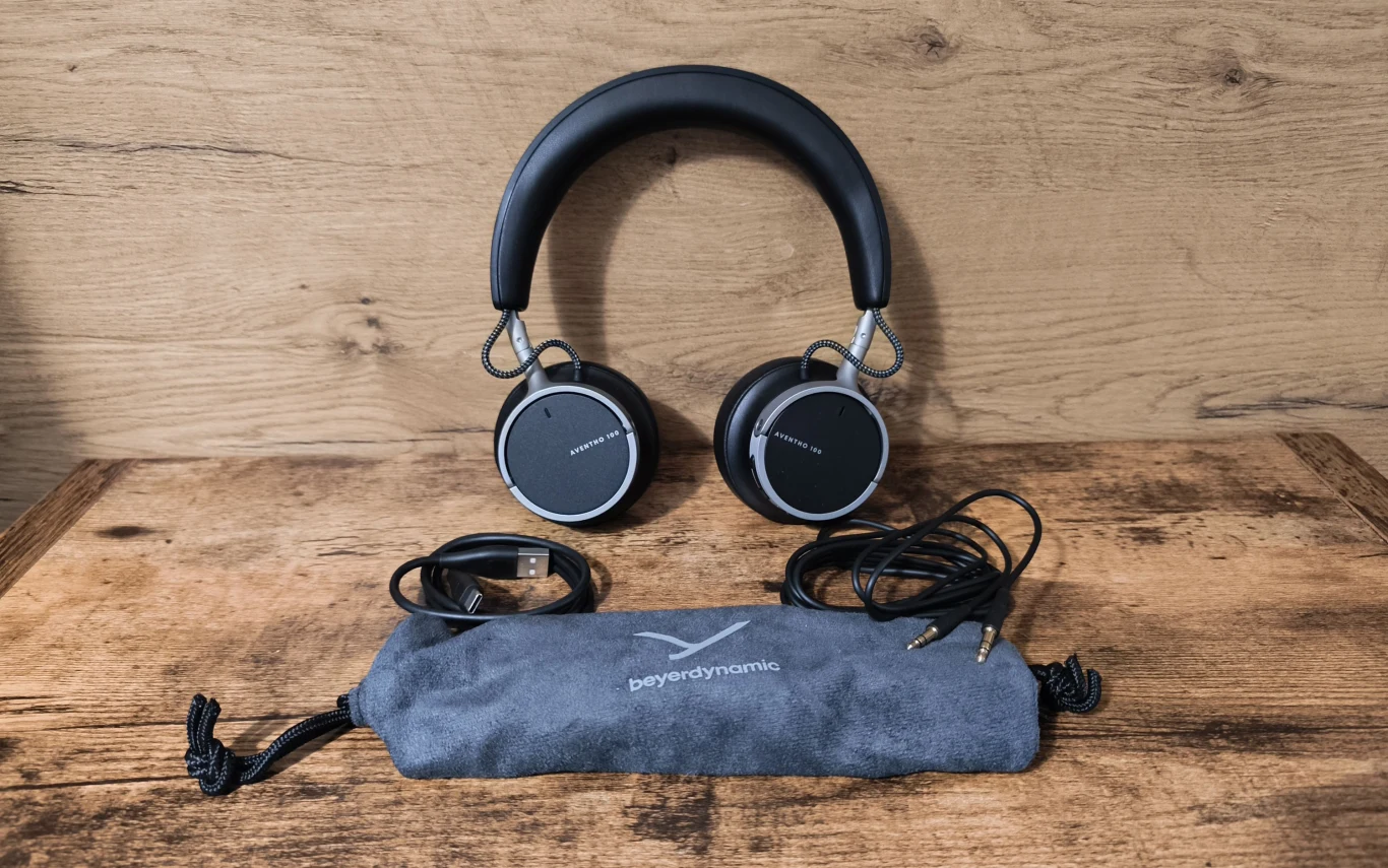 Beyerdynamic Aventho 100 Bezprzewodowe słuchawki nauszne marki Beyerdynamic w czarnym kolorze, kabel audio, kabel USB oraz szara materiałowa saszetka z logotypem producenta ułożone na drewnianej powierzchni.