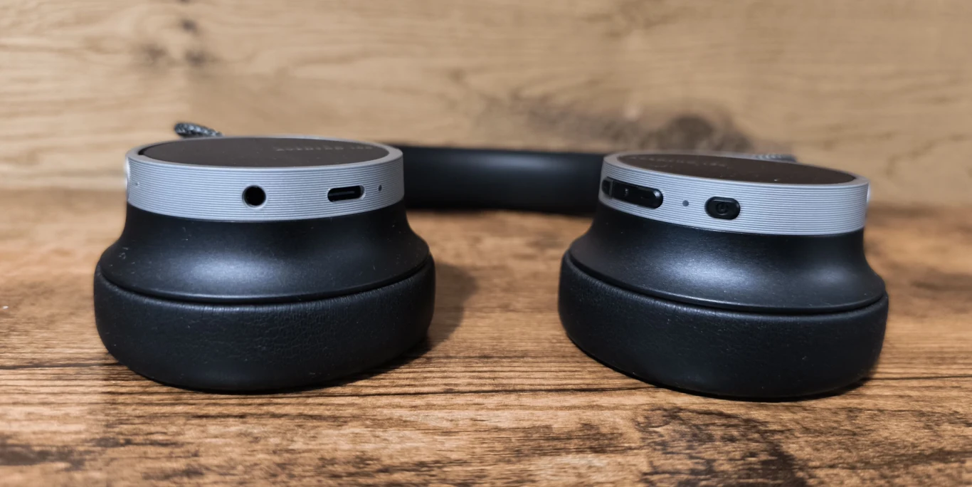 Beyerdynamic Aventho 100 Czarne nauszniki słuchawek bezprzewodowych z widocznymi srebrnymi elementami oraz portem ładowania USB i przyciskami sterującymi, ułożone na drewnianej powierzchni.
