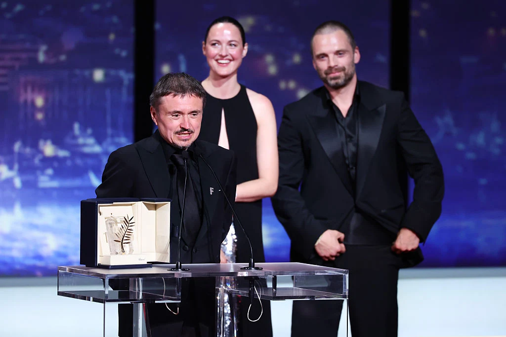Mężczyzna w ciemnym garniturze przemawia przy mikrofonie, na przezroczystym podium stoi prestiżowa nagroda w ozdobnym pudełku, za nim uśmiechnięta kobieta i towarzyszący jej mężczyzna również ubrani elegancko.