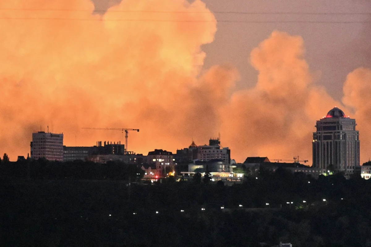 Panorama miasta z wyraźnie widoczną ścianą jasnopomarańczowego dymu unoszącego się nad budynkami, w tym kilkoma wysokimi wieżowcami i żurawiem budowlanym, podkreślająca atmosferę niepokoju.