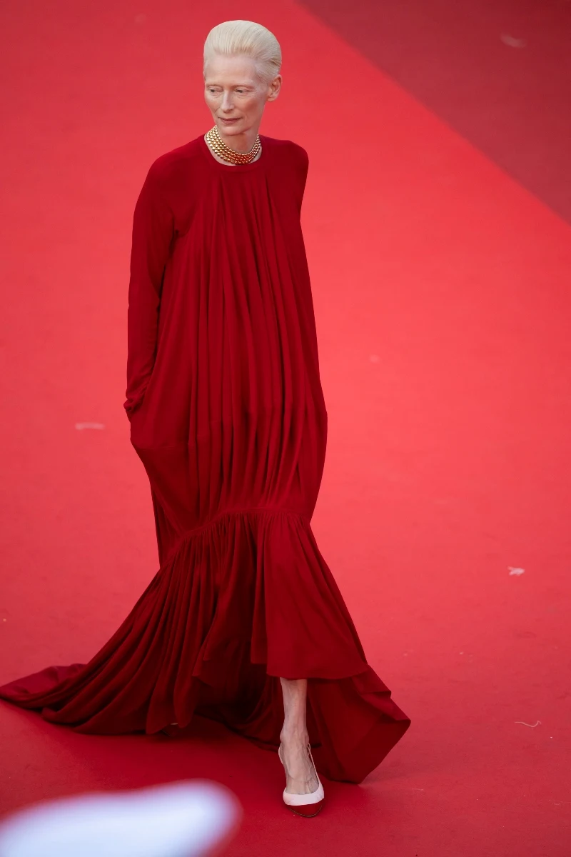 Tilda Swinton w Cannes Osoba o jasnych włosach w długiej, czerwonej sukni z szerokim dołem idzie po czerwonym dywanie, nosząc jasne buty i złoty naszyjnik.