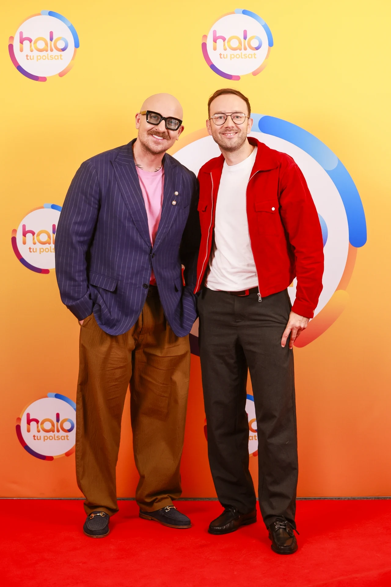 Robert Czerwik, Marcin Radomski Dwóch uśmiechniętych mężczyzn stoi na czerwonym dywanie przed kolorową ścianką z logotypami „Halo tu Polsat”; jeden ubrany w niebieską marynarkę i brązowe spodnie, drugi w czerwoną kurtkę i czarne spodnie.