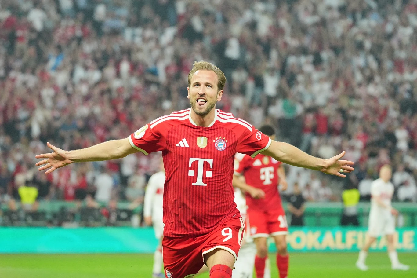 Piłkarz Bayernu Monachium w czerwonym stroju z rozłożonymi rękami świętuje zdobycie bramki na tle kibiców na stadionie.