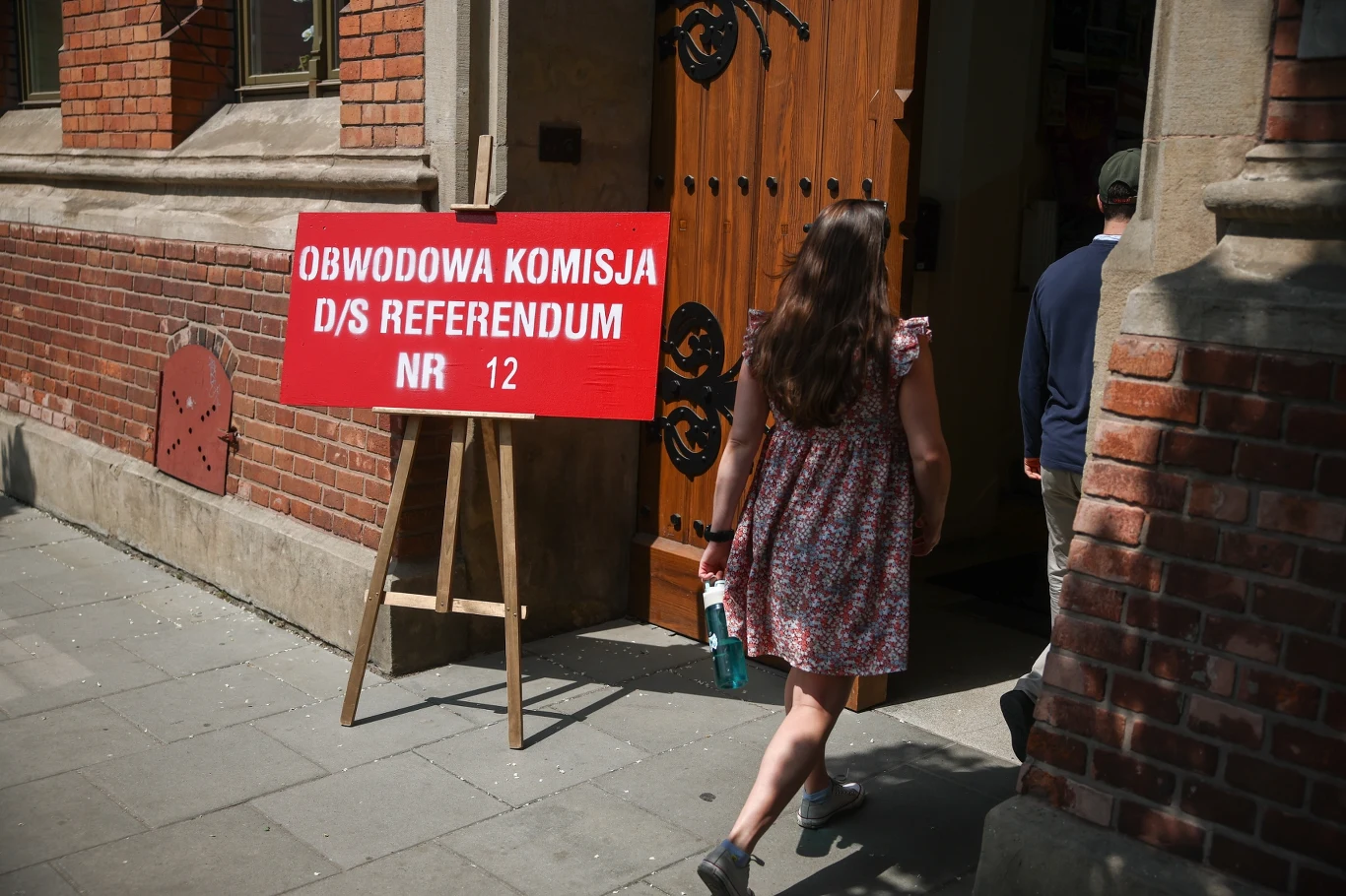 Komisje referendalne w Krakowie są odpowiednio oznakowane Czerwona tablica z napisem 'Obwodowa komisja d/s referendum nr 12' ustawiona na chodniku przed wejściem do budynku, do którego wchodzi kobieta w letniej sukience.