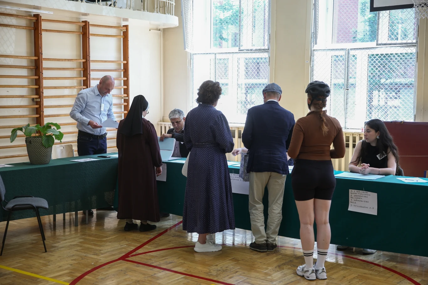 Referendum w Krakowie. Aby zagłosować trzeba pokazać członkowi komisji dokument tożsamości Grupa osób stoi przy stolikach komisji wyborczej w sali gimnastycznej, część z nich rozmawia z członkami komisji, a jedna osoba wypełnia dokumenty.