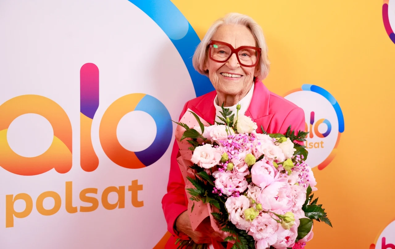 Teresa Godlewska w "halo tu polsat" Teresa Godlewska w różowym żakiecie z bukietem jasnoróżowych kwiatów na tle ścianki z logo Polsat.