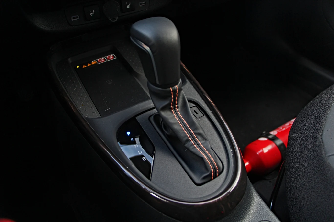 Jak każda hybryda Toyoty, także Aygo X korzysta z przekładni e-CVT Dźwignia automatycznej skrzyni biegów z widocznymi oznaczeniami pozycji oraz gaśnica umieszczona obok fotela kierowcy.