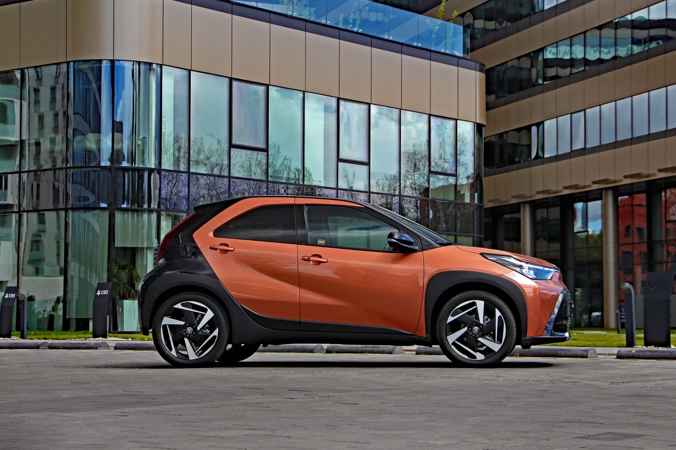 Zwis przedni został w Aygo X Hybrid wydłużony o 76 mm, ale zupełnie tego nie widać Pomarańczowo-czarny miejski samochód zaparkowany przed nowoczesnym biurowcem z dużymi, przeszklonymi fasadami.
