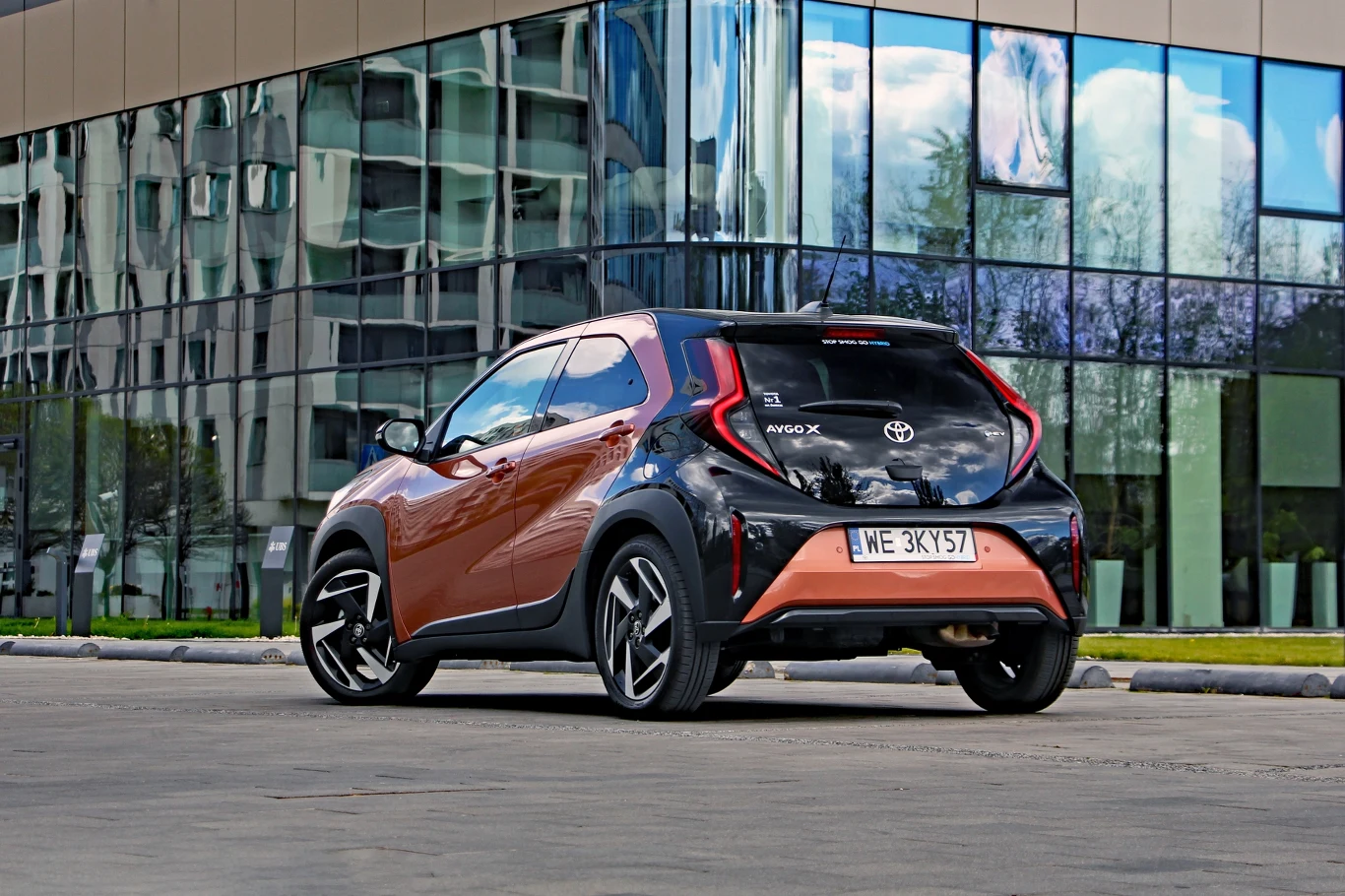 Dopiero spojrzenie na tył Aygo X Hybrid zdradza, że mamy do czynienia z wciąż tą samą generacją modelu Pomarańczowo-czarna Toyota Aygo X stoi zaparkowana na tle nowoczesnego przeszklonego budynku, kontrastując swoim kompaktowym nadwoziem z geometryczną architekturą w odbiciu szyby.